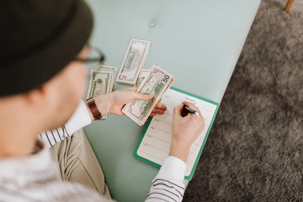 Person counting U.S. dollar bills and writing in a budget planner at a table.